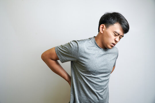 A young Asian man holding his lower back with both hands, with a grimacing expression of pain and discomfort.