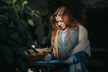 Focused woman in corporate attire takes notes on paper while outside surrounded by greenery,...
