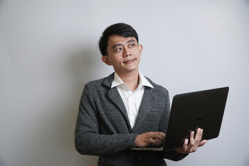 A thoughtful young Asian businessman in a grey suit holds a laptop while looking up and to the...