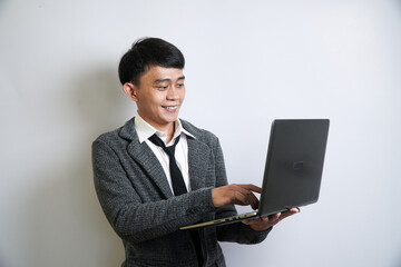 A happy young Asian businessman in a grey suit and loosened tie smiles while standing and working on a laptop he holds in his hands.