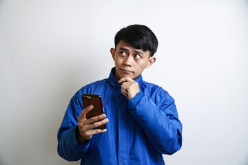 A young Asian man in a blue jacket holds a phone and looks away thoughtfully, with his hand on his...