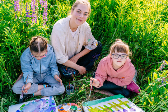 Inclusive outdoor art class with teacher and two girls painting in a wildflower meadow