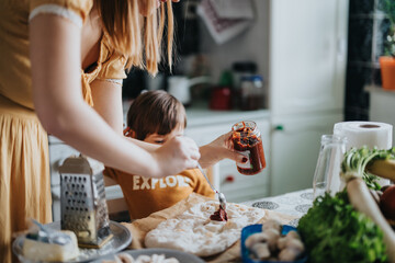 A woman and her child enthusiastically preparing a pizza together in a warm kitchen setting,...
