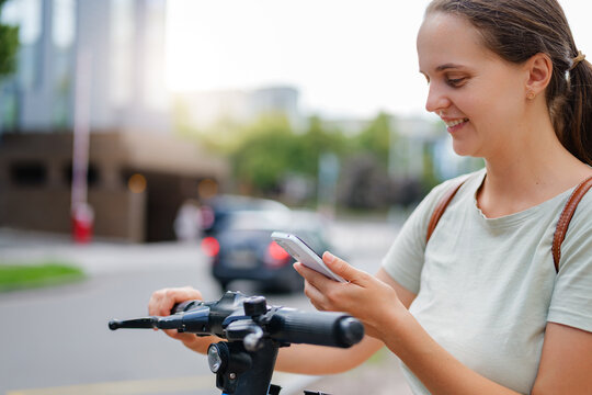 A happy young woman on a city street smiles while using her smartphone to interact with an electric scooter, blending technology with urban mobility.