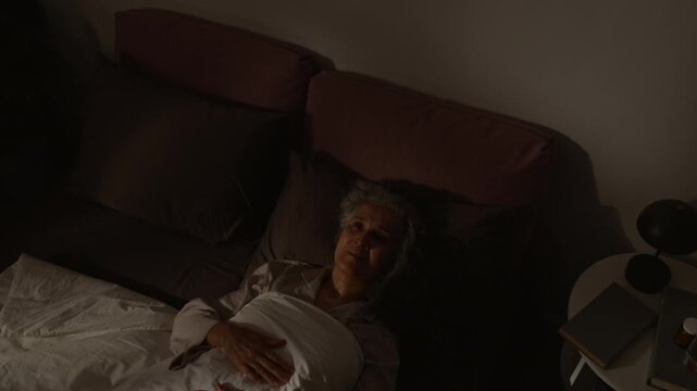 Medium close-up of tearful senior Caucasian woman with gray hair lying alone in bed with open eyes, wide awake, staring into darkness, unable to sleep, suffering from insomnia or depression