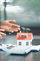 Real estate agent and customer are discussing contract to buy house, with house model in background symbolizing real estate, insurance, and loan arrangements involved in process.
