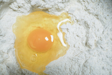 Cracked egg rests in mound of white flour, ready for baking. yolk is vibrant and egg white is slightly spread, creating contrast with flour. This setup is perfect for making dough or batter