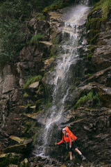 a beautiful girl with a hiking backpack is standing by a waterfall in a forest in the mountains. a female traveler exploring the forest. the concept of travel and wanderings. summer vacation, a place 
