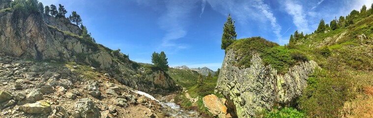 Panoramic mountain landscape with rocky terrain, trees, and a clear sunny sky