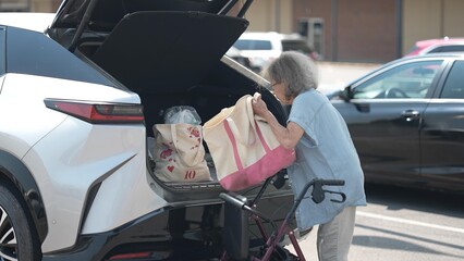 An elderly woman is grocery shopping while using a walker. She is loading her bags into the trunk...