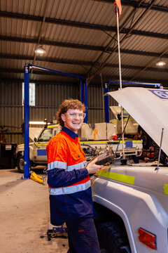 Young mechanic inspecting mining fleet vehicle inside industrial workshop holding diagnostics device