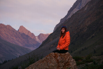 Obraz premium A young woman is sitting on a large rock and enjoying the view of the mountains. Hiker in the mountains during sunset, outdoor activities