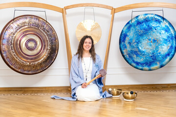 Woman in ceremonial robe smiling while sitting between colorful gongs with singing bowls on the...