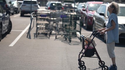 A disabled elderly woman uses her walker in a supermarket parking lot, carefully navigating her way...