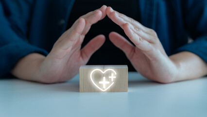 Hands protecting a wooden block with a glowing heart and plus sign icon, symbolizing health care,...