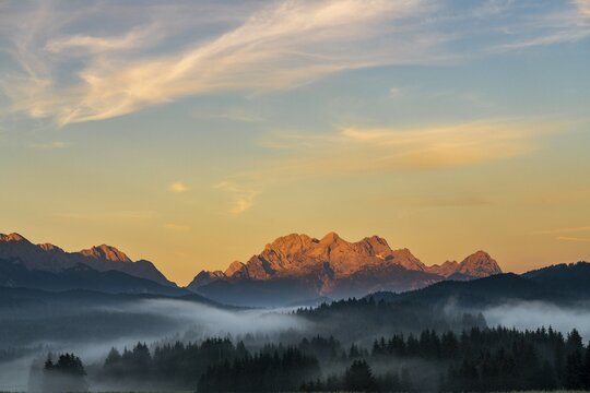 Mountain meadows with forest in light ground fog and Zugspitz massif in the background at sunrise, Kr&uuml;n, Bavaria, Germany