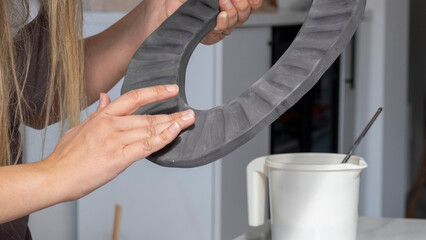 A close-up shot of a beautiful middle-aged woman painting a clay ceramic frame black at a table in an art studio, with artistic paintings adorning the background