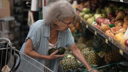 An elderly senior woman carefully chooses fresh avocados from a grocery store display filled with various fruits, showcasing her dedication to healthy eating.