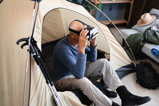 Senior man sitting in the camping tent having fun playing with virtual reality glasses at home indoors