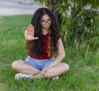 teenager with long black hair sitting cross legged on grass holding up hand in a stop gesture