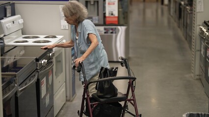 An elderly woman using a walker carefully examines kitchen appliances in a hardware store, focused...