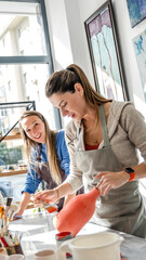 Obraz premium In an art studio with an environment adorned with artistic paintings in the background, two beautiful middle-aged women are joyfully painting a clay ceramic vase pink together at a table, having fun