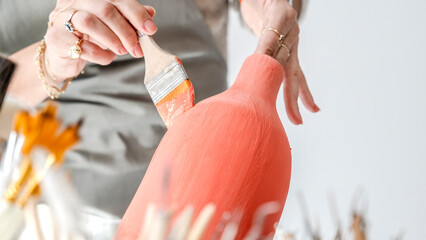 A close-up shot of a beautiful middle-aged woman painting a clay ceramic vase pink at a table in an art studio