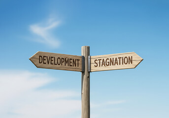 A wooden signpost with two arrows points to "DEVELOPMENT" and "STAGNATION" against a cloudy blue sky.