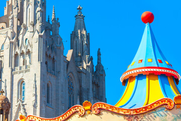 Fototapeta premium Colorful roof of carousel at Tibidabo Amusement Park with the Gothic style Temple of Sacred Heart in Barcelona in background. Blue sky contrasts with vibrant amusement park detail and stone church