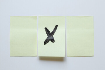 A yellow sticky note with a black watercolor cross on it, placed between two blank sticky notes, on...