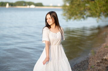 Young woman in long white dress standing on riverbank, enjoying summer sunset and fresh breeze.