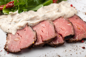 Fototapeta premium Sliced roast beef served with a creamy sauce and fresh arugula on a white plate