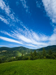 Obraz premium A vibrant green meadow foregrounds a forested hill under a bright blue sky with wispy clouds, evoking peace and nature, suitable for wellness, travel, and eco-friendly promotions.