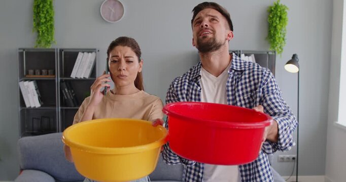 Worried couple catching water from a leaking ceiling with plastic buckets while the woman calls for help. Concept of apartment problems, water damage, and emergency repair situations. 