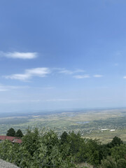 Wide panoramic view of a rural valley with green fields, scattered houses, and a bright blue sky...