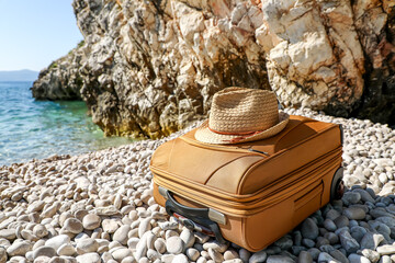 Yellow suitcase with a straw hat on top stands on a rocky beach, with turquoise sea waves and steep...