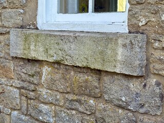 Close-up of an old stone windowsill beneath a white wooden-framed window. The weathered stone and textured masonry wall show signs of age, moss growth, and natural discoloration.