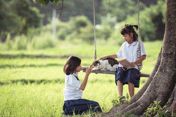 Two Girls Sitting on Swing in a Green Field with a Cat, Enjoying a Quiet Moment in Nature Surrounded by Lush Grass and Trees in Bright Daylight