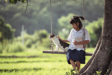 Young girl in school uniform enjoying a serene moment with a playful cat on a swing in a lush green field under a tree in the afternoon light