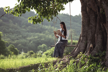 Woman Reading Book on Swing Under Large Tree in Scenic Green Landscape Surrounded by Nature and Mountains During Sunny Day