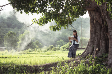 Young woman reading a book while sitting on a swing under a large tree in a serene rice field landscape, surrounded by lush green nature and soft mist