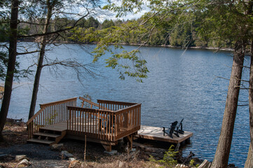 A wooden dock and deck on a calm blue sunlit lake surrounded by forest. Adirondack chairs are on...