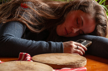 A close-up of a woman with her head down on a rustic wooden table, with her eyes closed, and her phone held in her hand, suggesting exhaustion or rest.