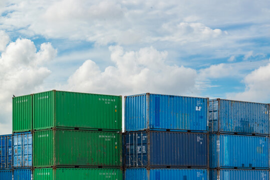 Green and blue shipping containers stacked at a port under a cloudy blue sky, representing global trade, cargo transport, and logistics.