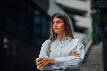 Businesswoman holding takeaway coffee is waiting with crossed arms in front of modern office building