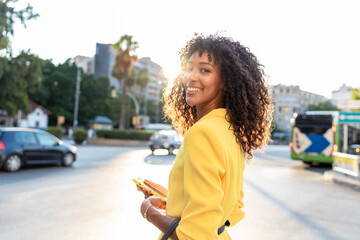 African-American woman in yellow blazer holding smartphone and smiling while walking in city at sunset. Concept of business, lifestyle, confidence.