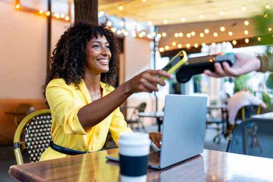African-American woman in yellow blazer using smartphone for contactless payment at café table with laptop. Concept of business, technology, mobile payment. - Powered by Adobe