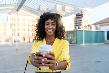 African-American woman in yellow blazer holding and offering a takeaway coffee cup outdoors....