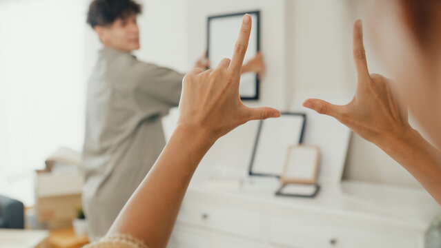 Happy Asian couple help each other decorate new space put photo frame on wall together in living room at new house. Husband and wife moving new house, Couple goal, Apartment rental and Owner Concept.