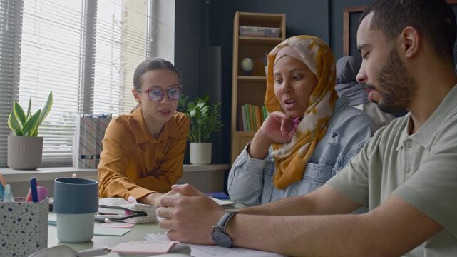 Multiethnic group of students and Caucasian female teacher working together at table during English class, discussing study materials in bright, well-organized classroom